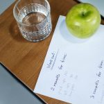 A green apple, glass of water, and workout plan on a wooden desk symbolize healthy living.